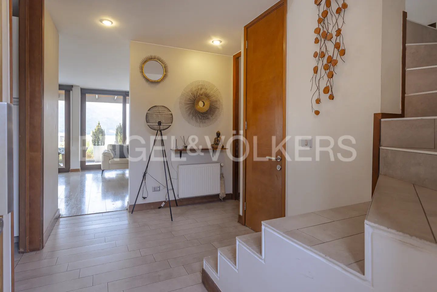 A bright foyer with tile floors, a wood door, and stairs. Wall decor includes a mirror, metal art, and a light fixture. A living room is visible in the background.