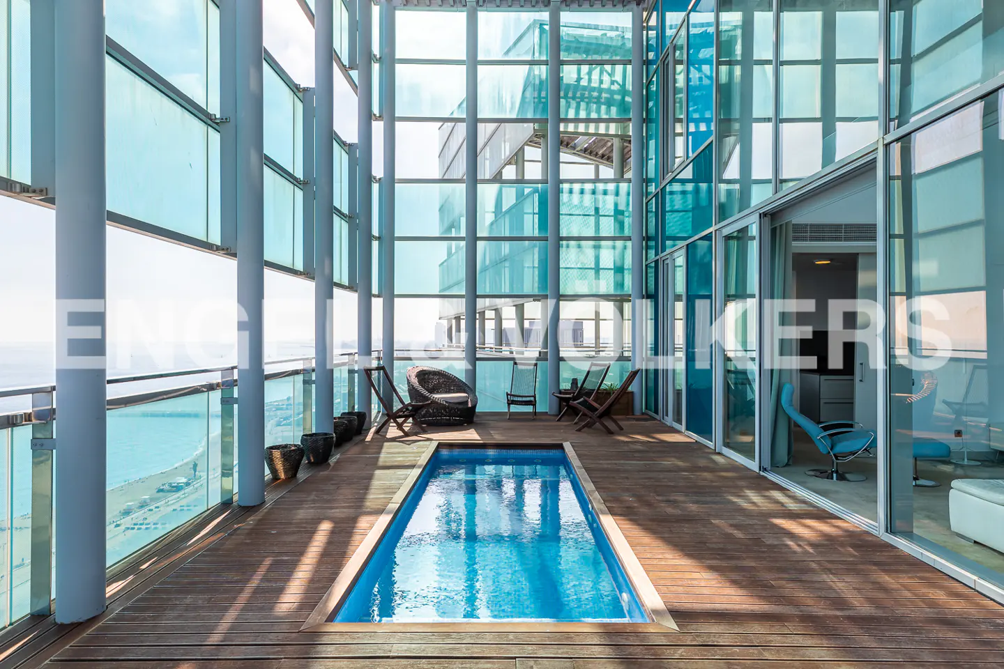 Modern penthouse balcony with a small pool, wood deck, and glass walls overlooking the ocean. Chairs are arranged for lounging.