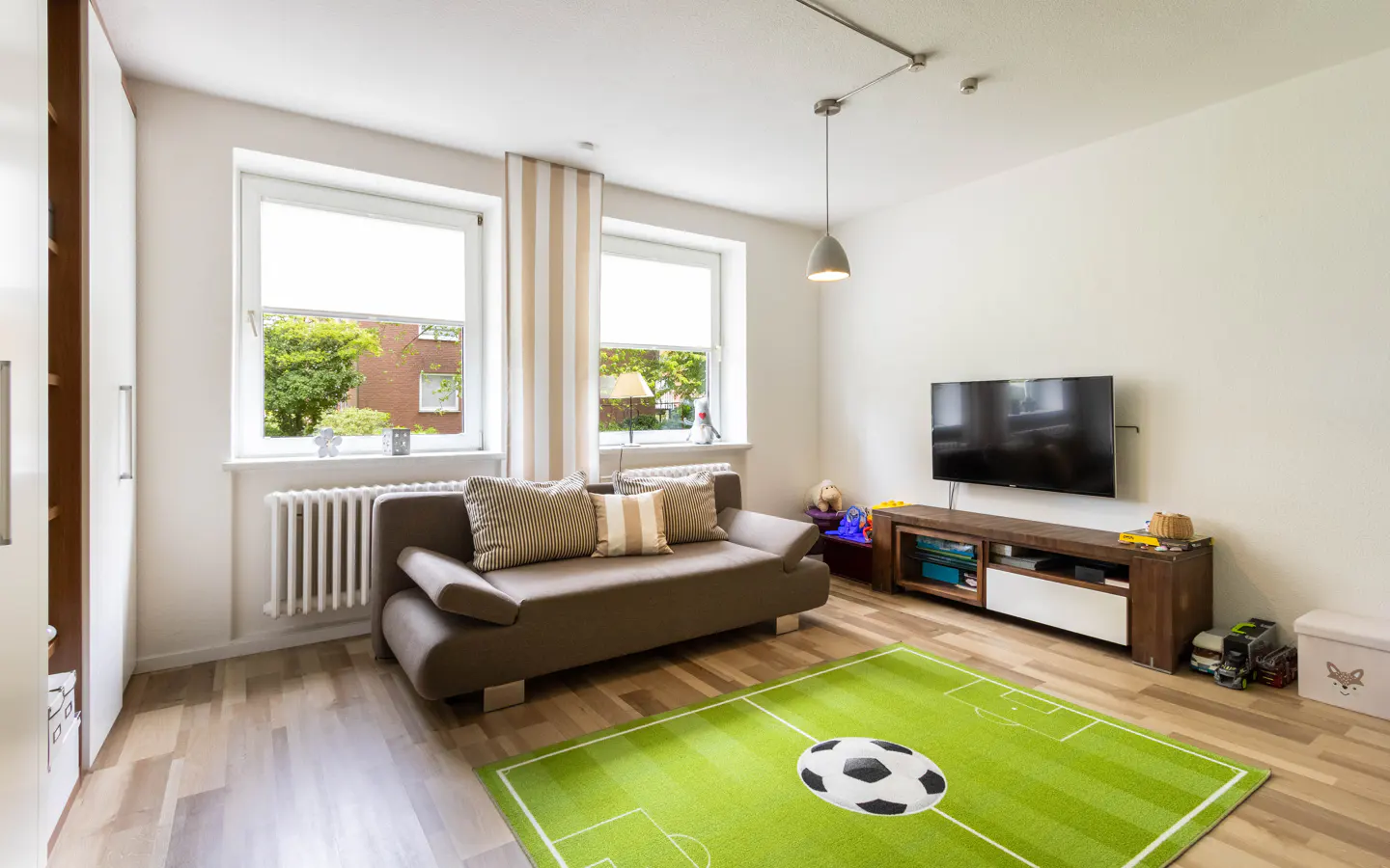A child's room with a brown sofa, TV, and a green rug with a soccer ball design. Two windows with white blinds let in natural light.