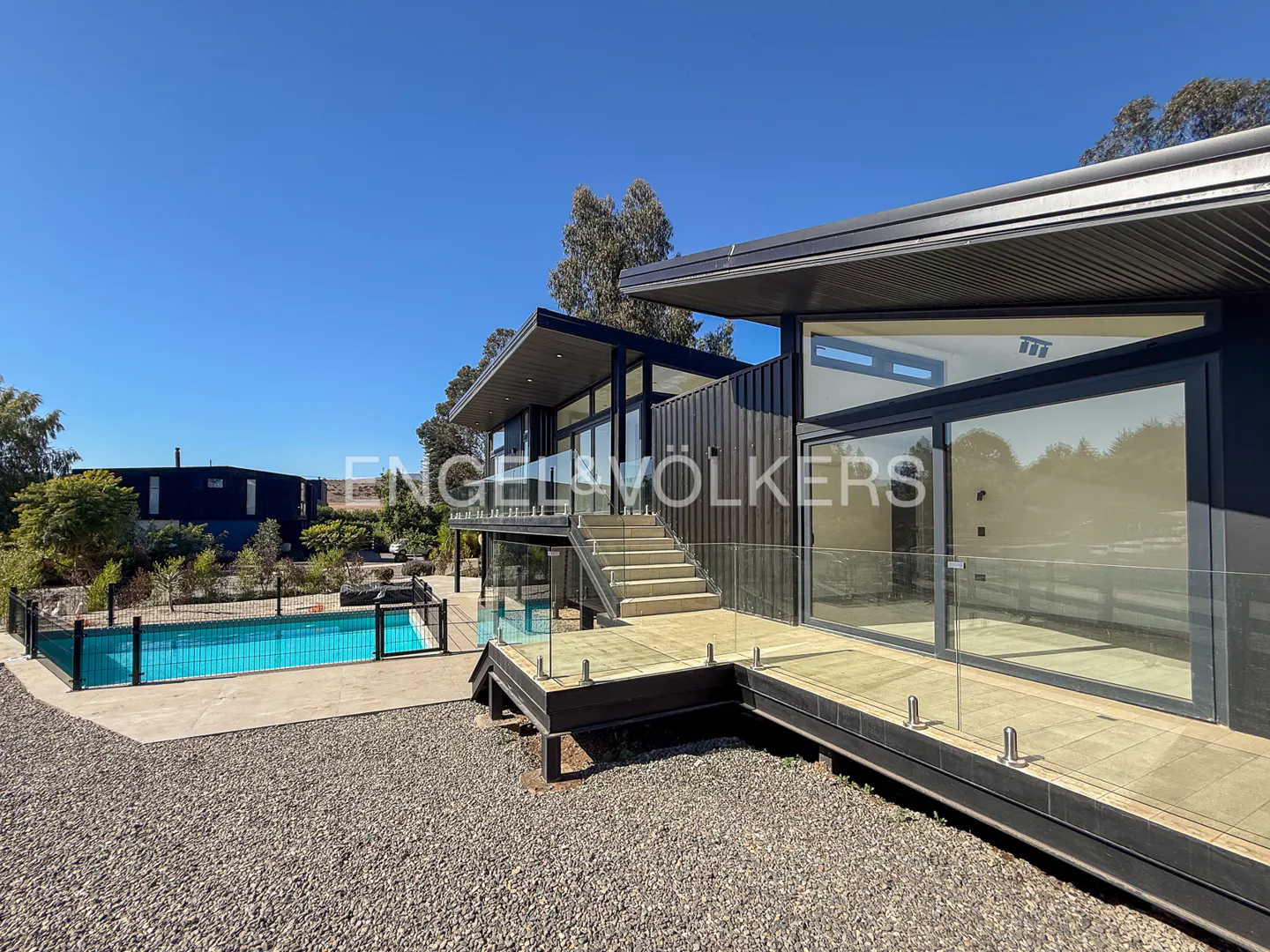 Modern black house with a wooden deck and glass railings, a pool, and a gravel yard under a clear blue sky.