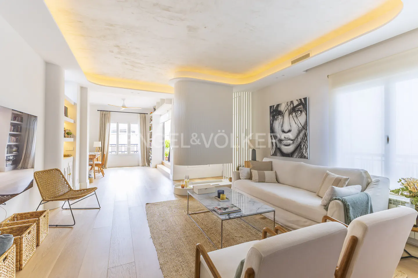 Bright, modern living room with white walls, light wood floors, and recessed lighting. White sofas, marble coffee table, and woven rug. Large portrait on the wall.