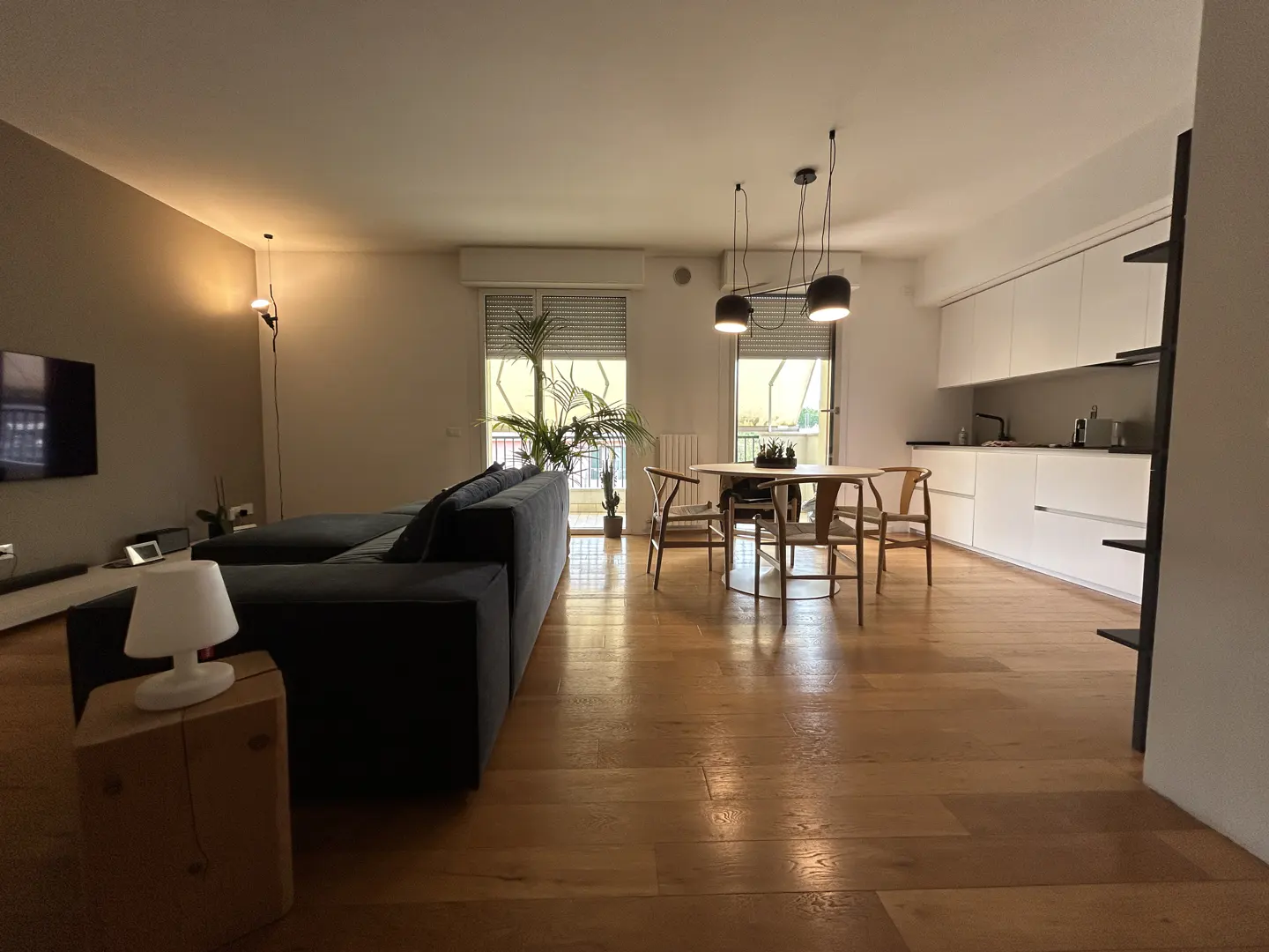 A modern apartment interior with a gray sofa, wooden floors, a round dining table, and a white kitchen.