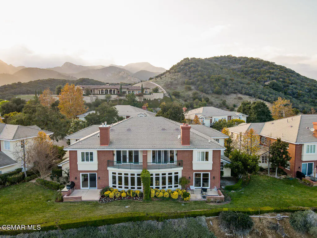 Aerial view of a large, red brick house with a tile roof, surrounded by green lawns and yellow flowers, with mountains in the background.