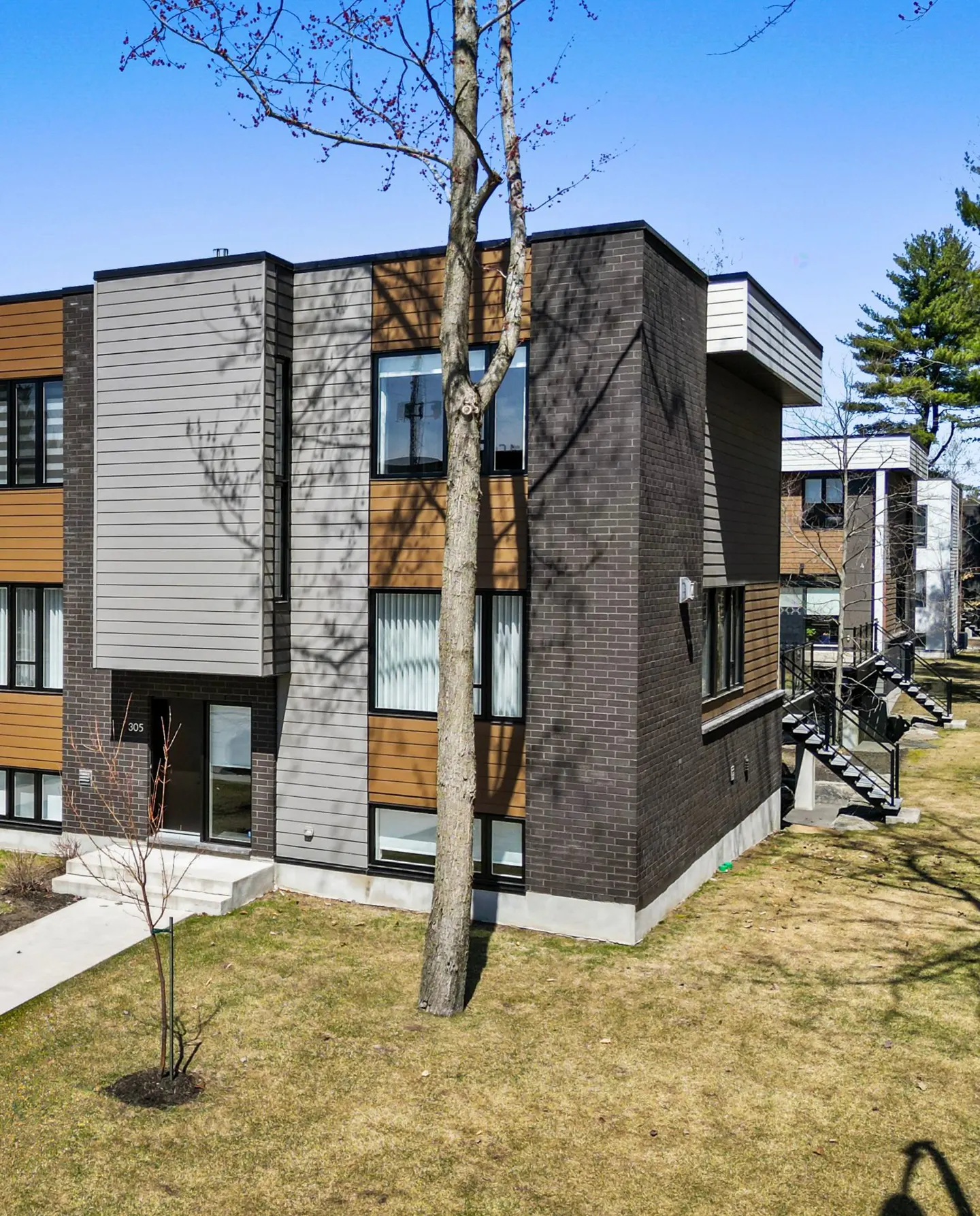 Modern apartment building with brown brick, gray siding, and wood-look panels, set on a grassy lawn under a blue sky.
