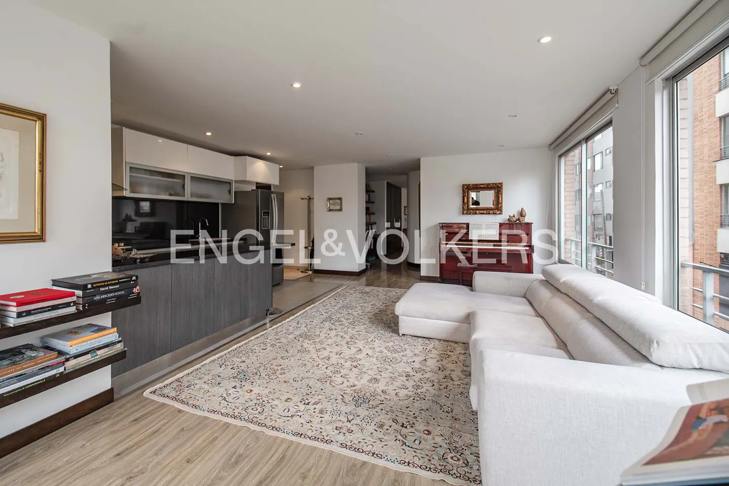 Open-concept living space with a white sofa, piano, and kitchen area. Large windows offer city views. A patterned rug covers the floor.