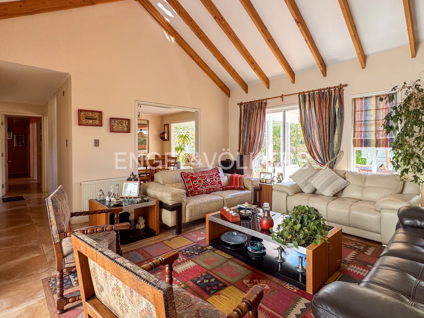 Living room with vaulted ceiling, exposed wood beams, and colorful rug. Beige sofas and wooden furniture fill the space.