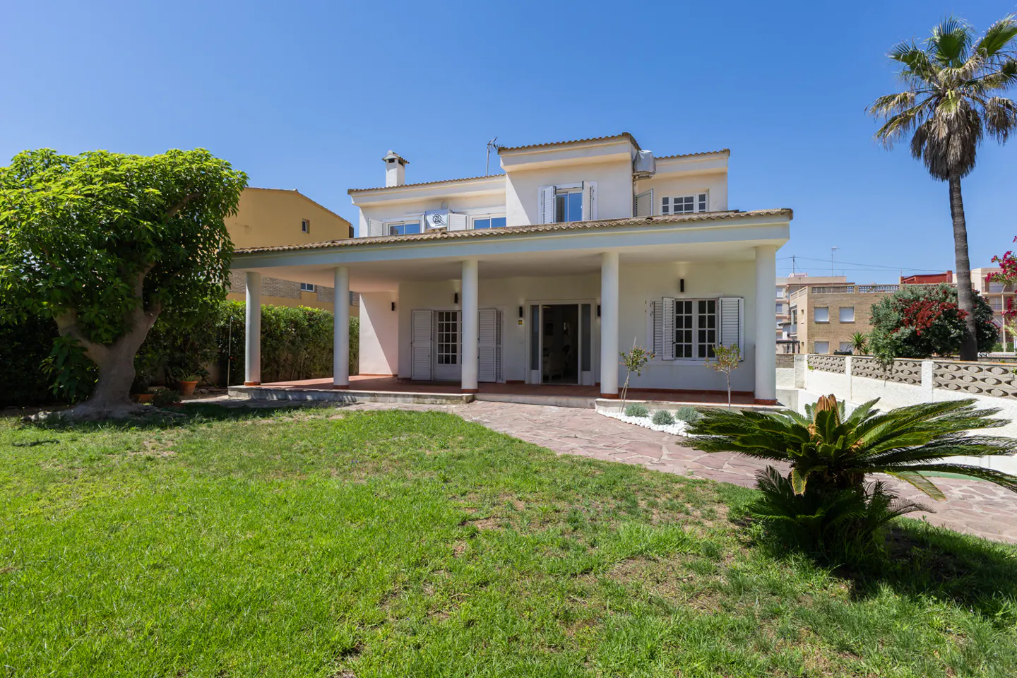Two-story cream villa with a large covered porch, white columns, green lawn, and palm tree under a clear blue sky.