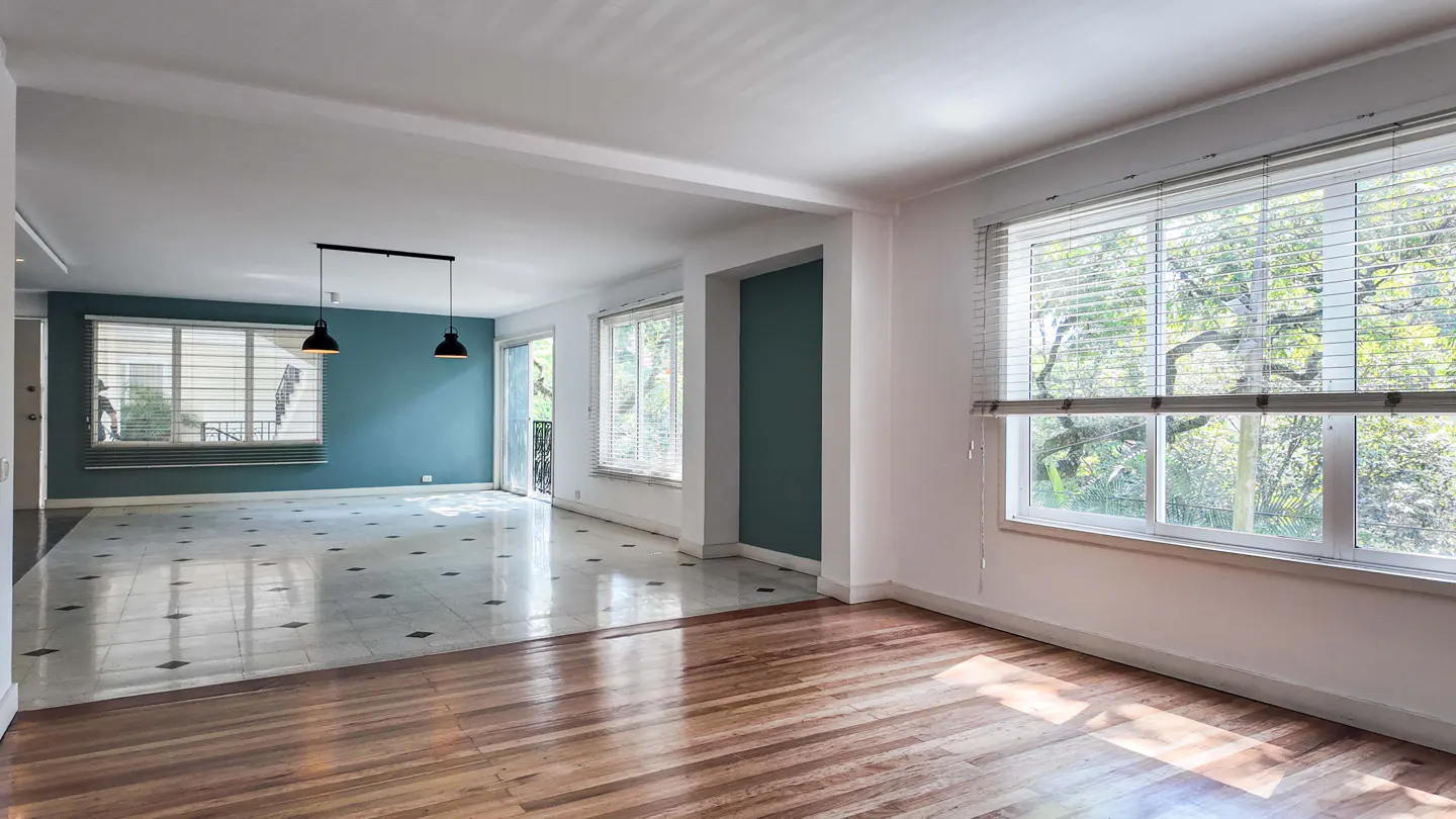 Bright, empty room with hardwood floors transitioning to tiled area. Large windows with blinds offer a view of lush greenery.