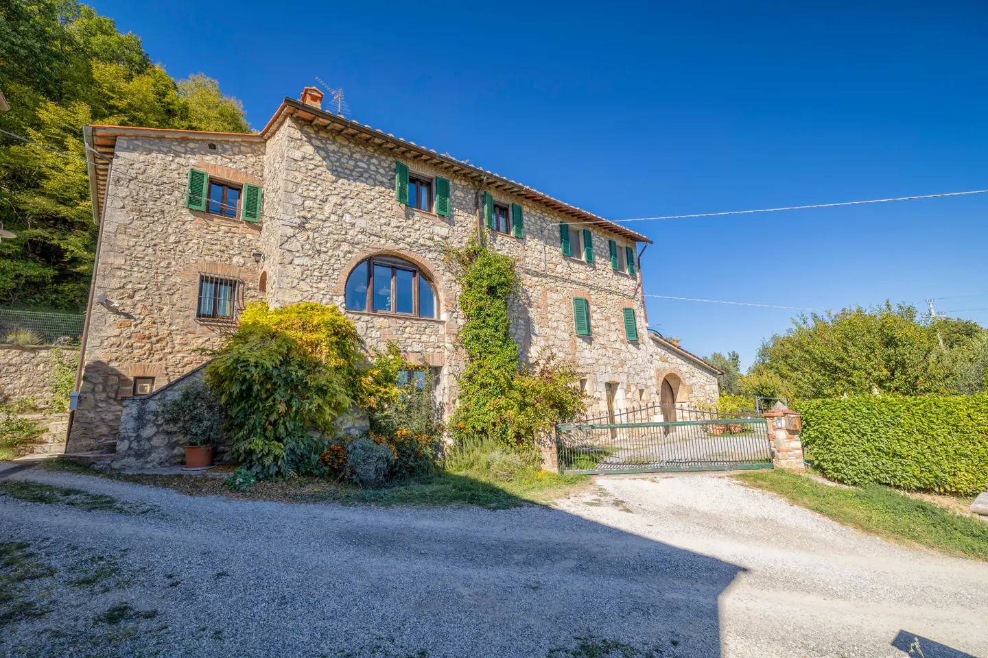 Exterior view of a two-story stone house with green shutters, a gated driveway, and lush greenery against a clear blue sky.
