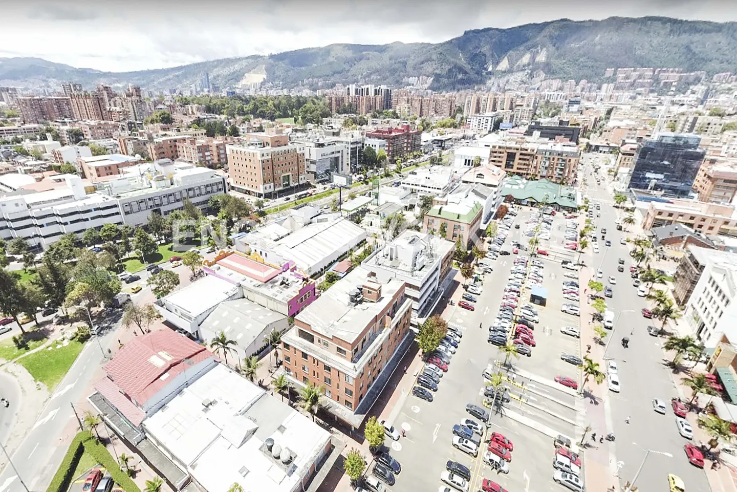 Aerial view of a city with buildings, trees, and cars, with mountains in the background.