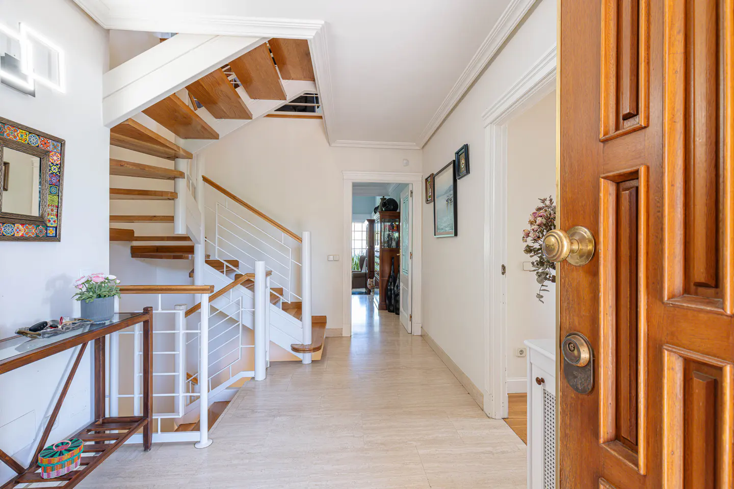 Open wooden front door reveals a bright foyer with stairs, a console table, and a hallway leading to other rooms.
