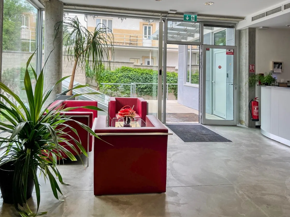 Lobby area with red chairs, glass table, and plants. Outside view through glass walls and doors. Reception desk and fire extinguisher visible.