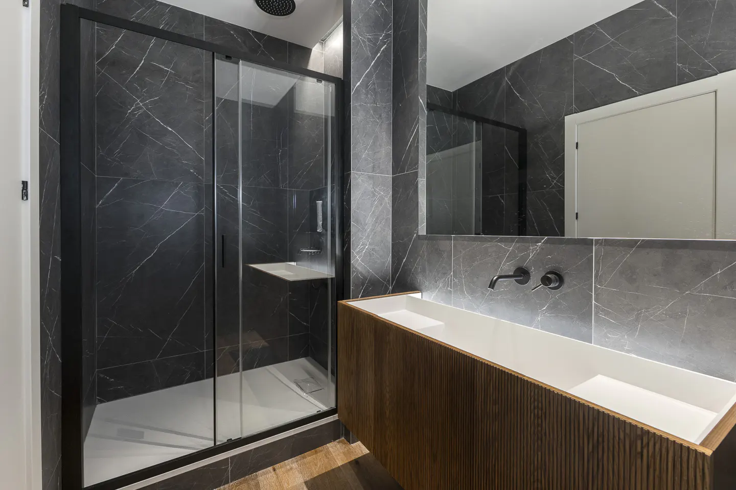 Modern bathroom with black marble walls, wood vanity with white sink, and glass shower with black frame.