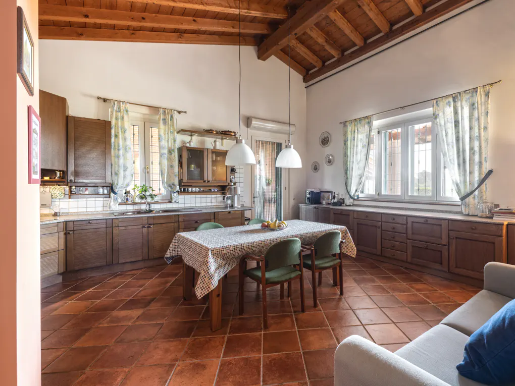 Rustic kitchen with wooden cabinets, terracotta tile floors, a dining table with green chairs, and exposed wood beams.
