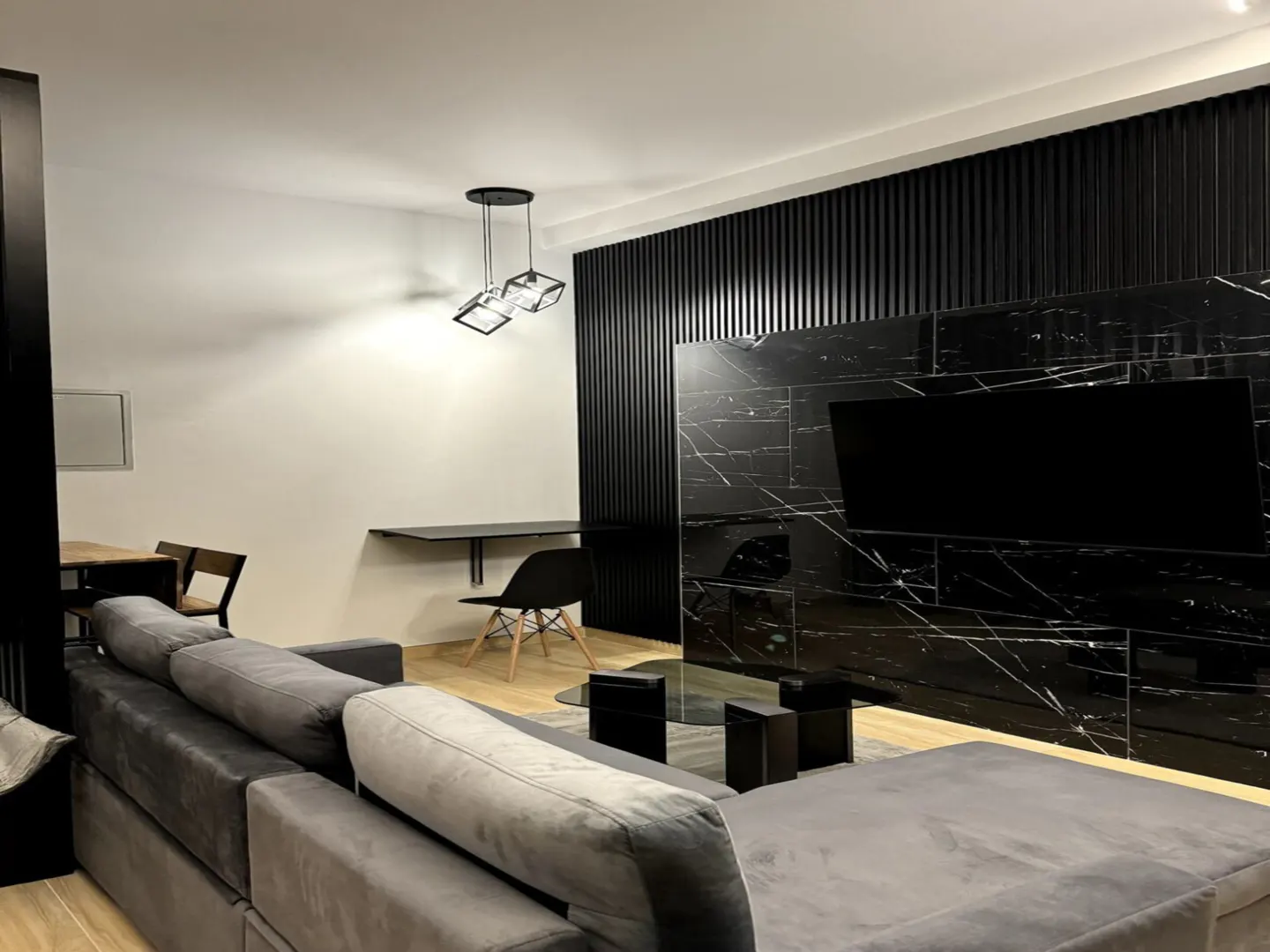 Modern living room with gray sectional sofa, black marble wall with TV, and black desk with chair. A geometric pendant light hangs above.