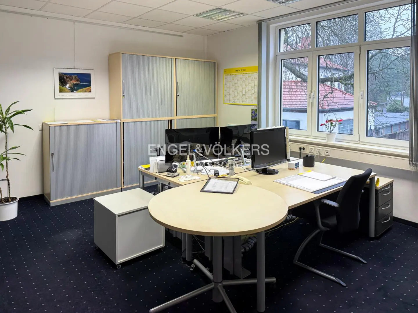 Office space with a round table, desk with monitors, filing cabinets, and a window overlooking a residential area. Engel & Völkers logo on the monitors.