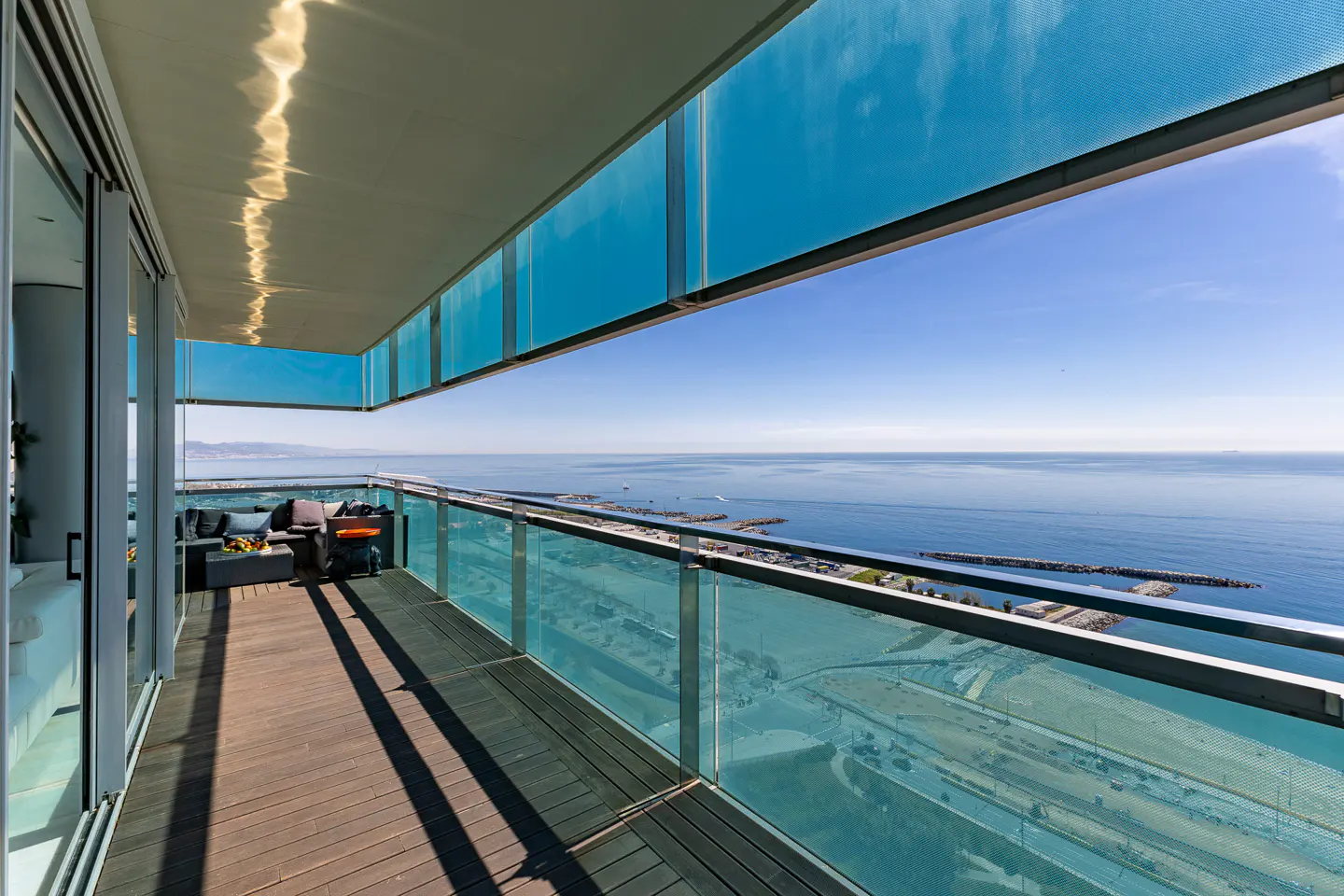 A modern balcony with a wood floor, glass railings, and ocean views. Outdoor furniture and a grill are visible.