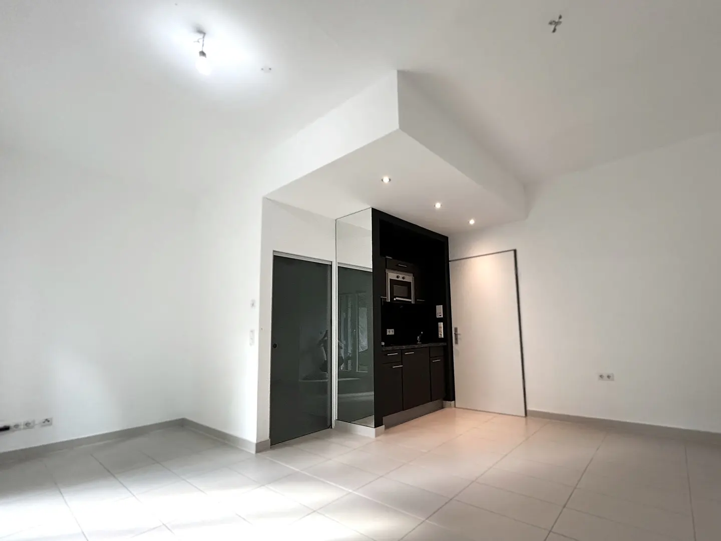 Bright, empty room with white walls and tile floor. A black kitchenette with microwave is on the back wall, next to a sliding door.