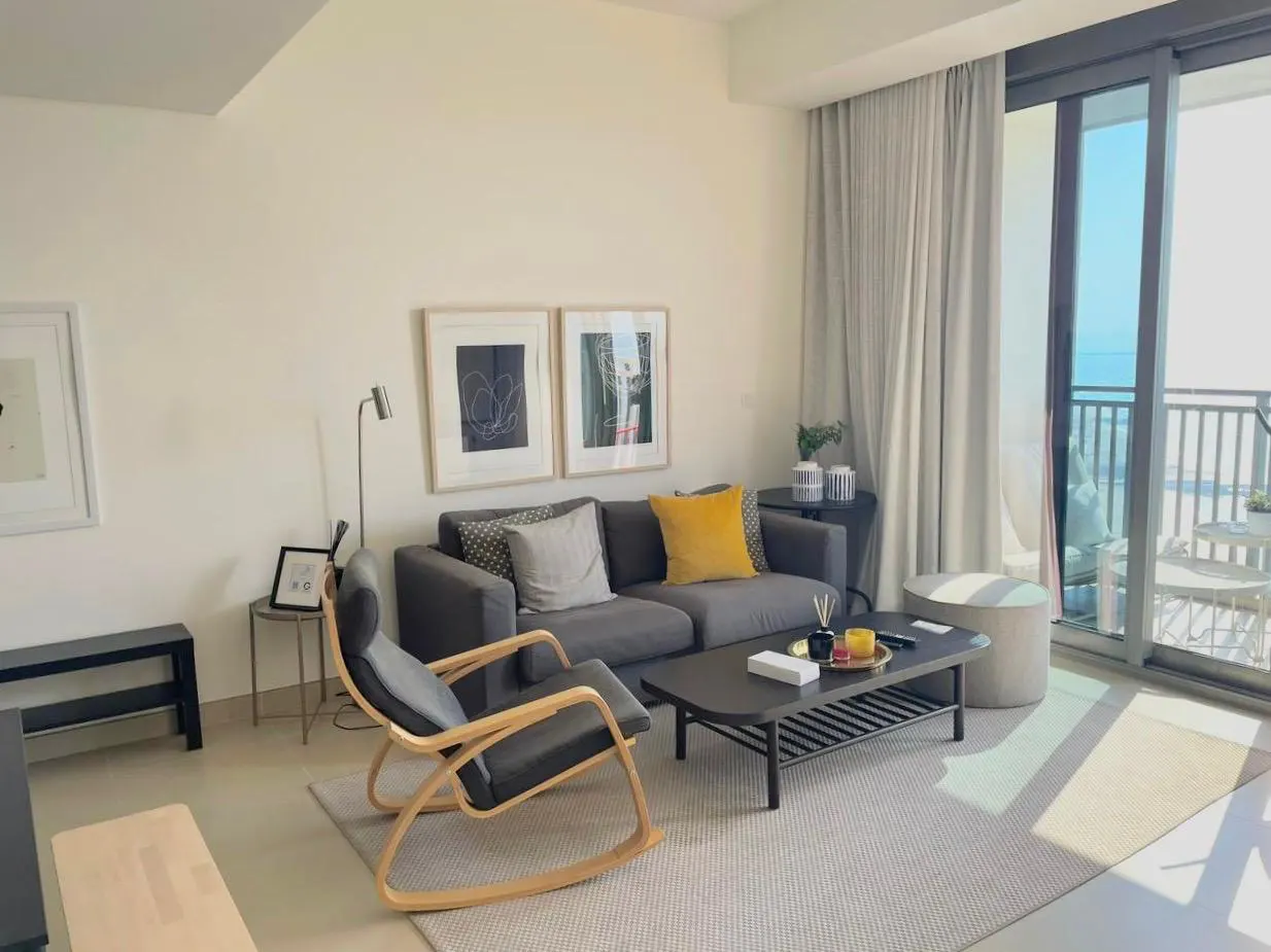 Living room with gray sofa, rocking chair, and coffee table on a gray rug. Balcony with ocean view in the background.