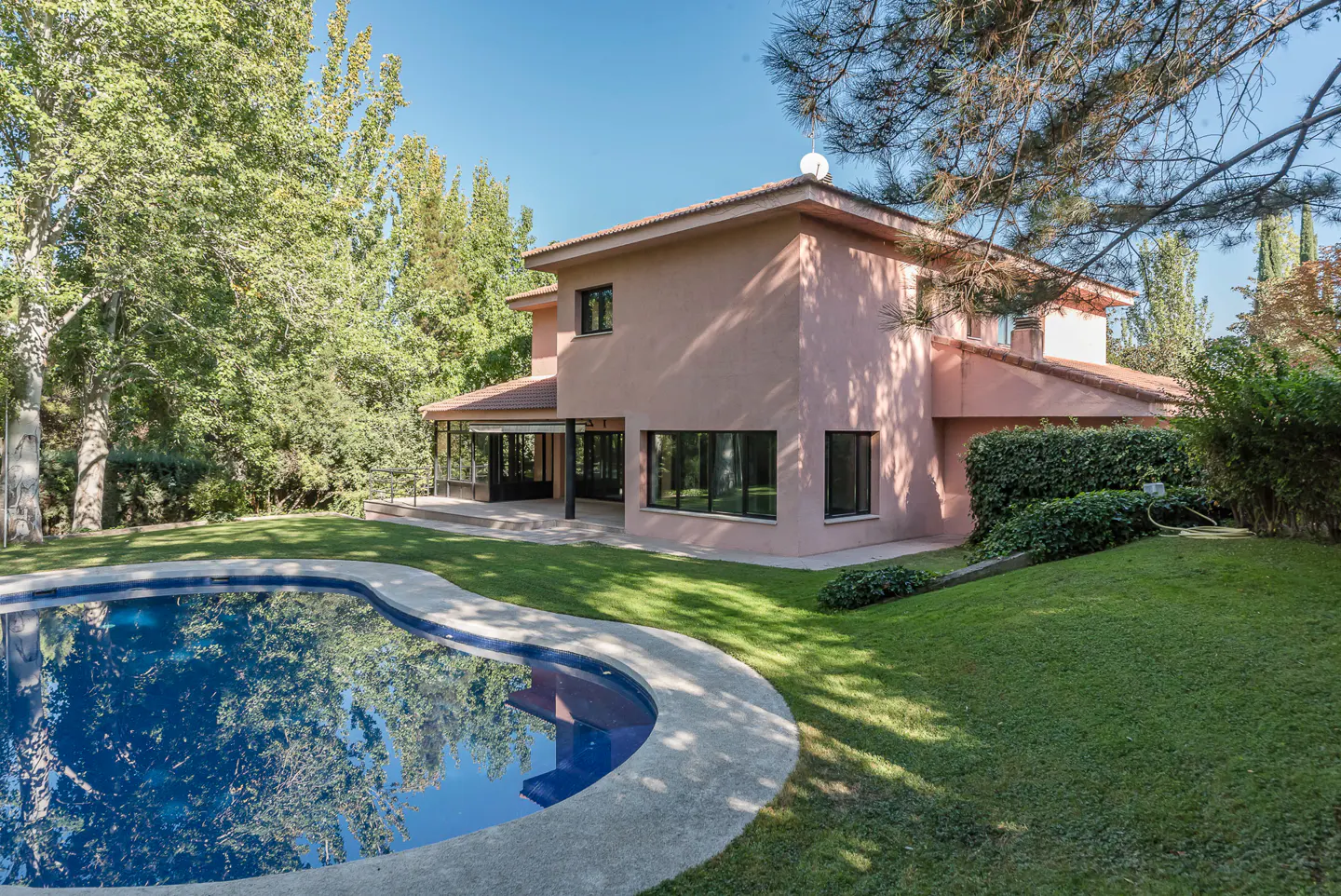 Two-story peach house with a red tile roof, a blue pool, and green trees under a blue sky.