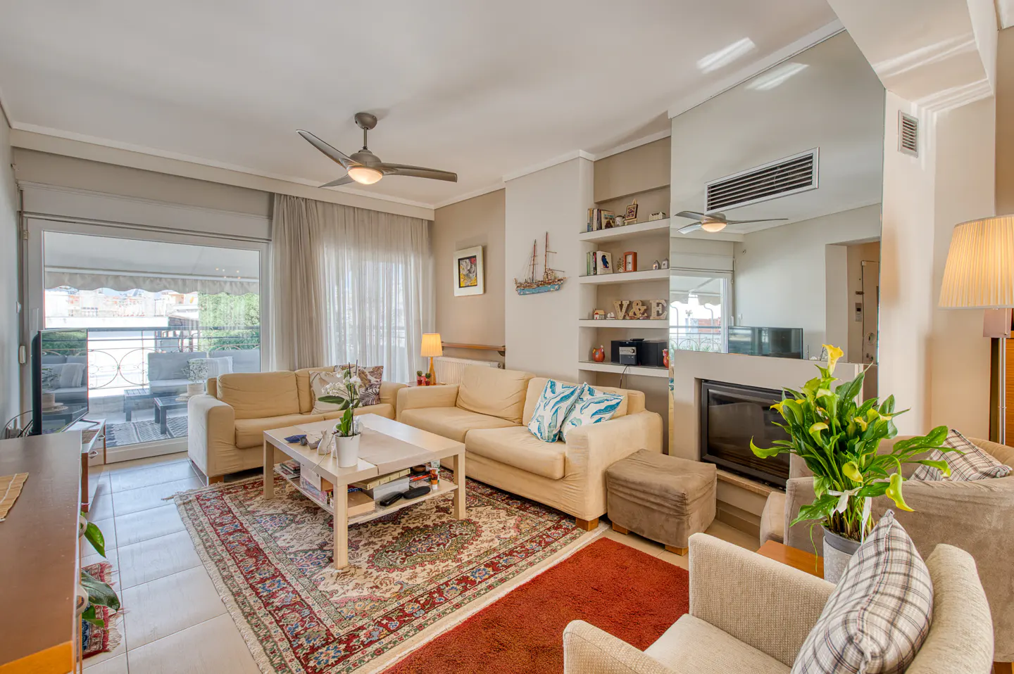 Living room with beige sofas, a patterned rug, and a fireplace. Shelves display decorative items. A ceiling fan hangs above. Balcony visible through glass doors.