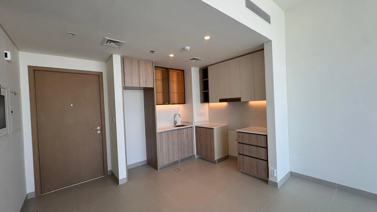 Modern kitchen area with light wood cabinets, white countertops, under-cabinet lighting, and a closed brown entry door.