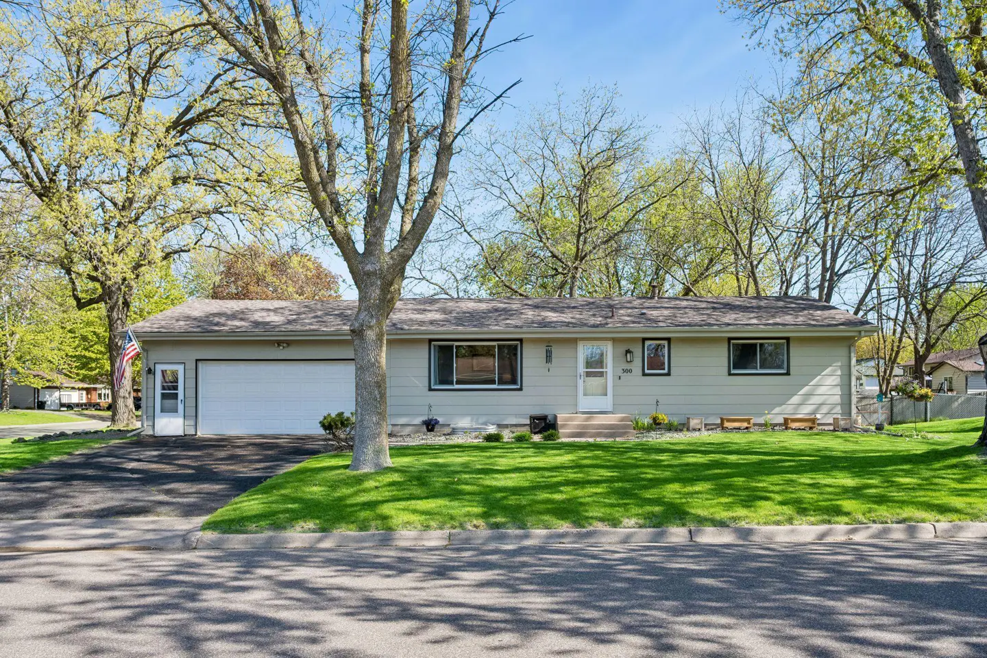 A single-story, light-green house with a white garage door and trim, set on a green lawn under a blue sky.