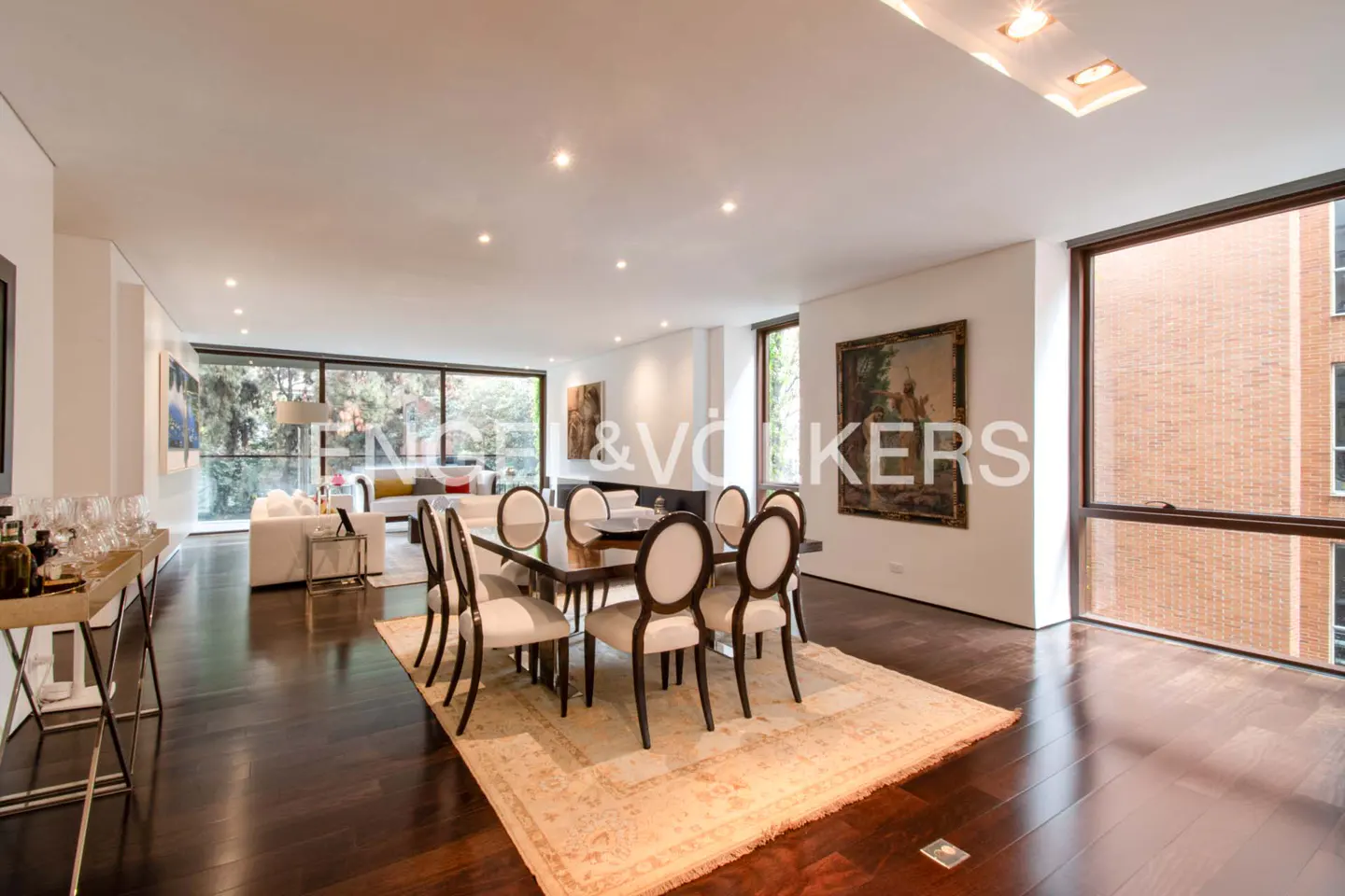 Bright, open-concept living and dining area with dark wood floors, a dining table with white chairs, and large windows.