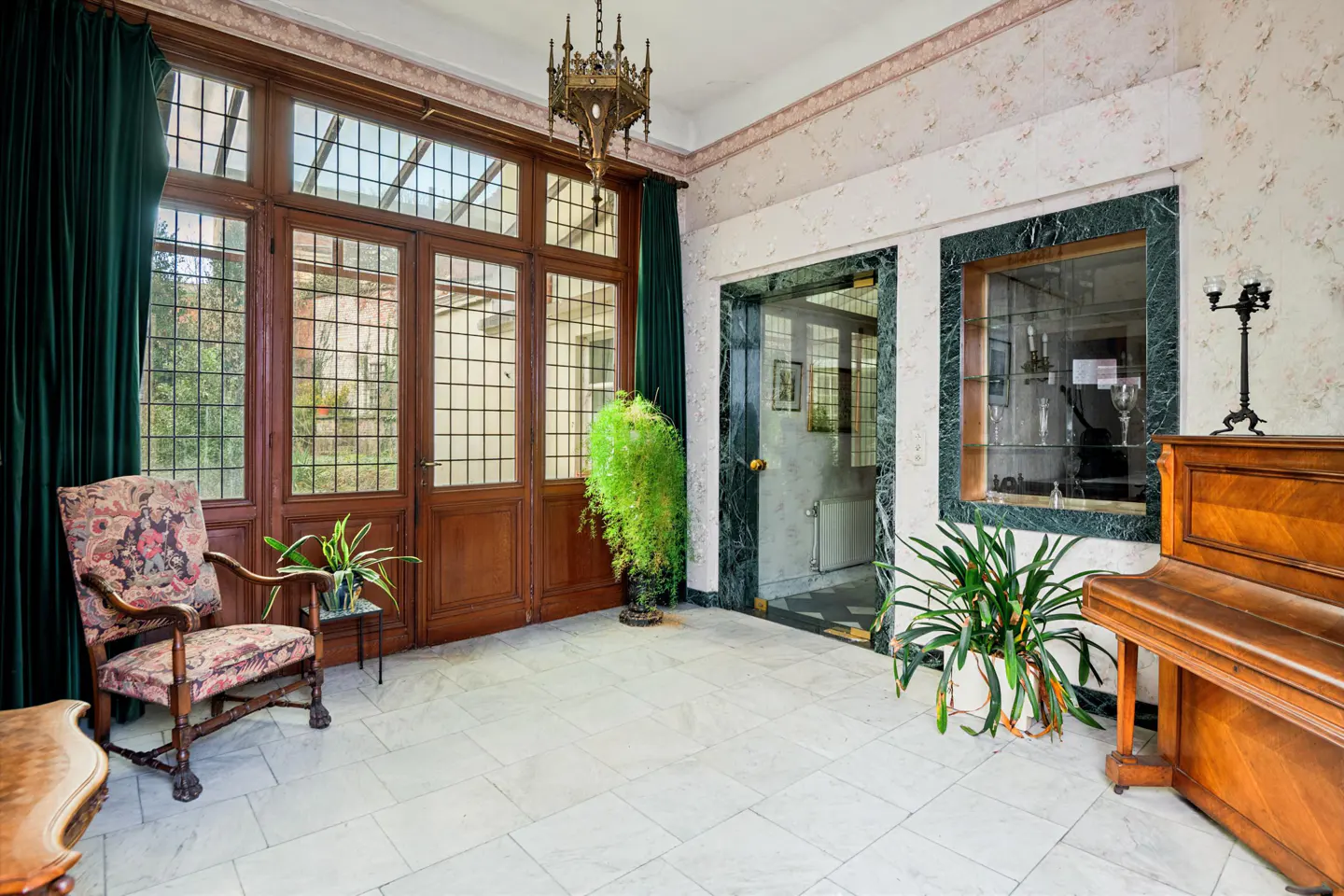 Sunroom with marble floors, wood-framed glass doors, and floral wallpaper. A piano and upholstered chair add to the room's charm.