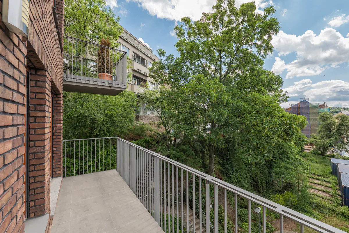Balcony view from a brick building, with gray railings, overlooking lush green trees and a distant building under a cloudy sky.