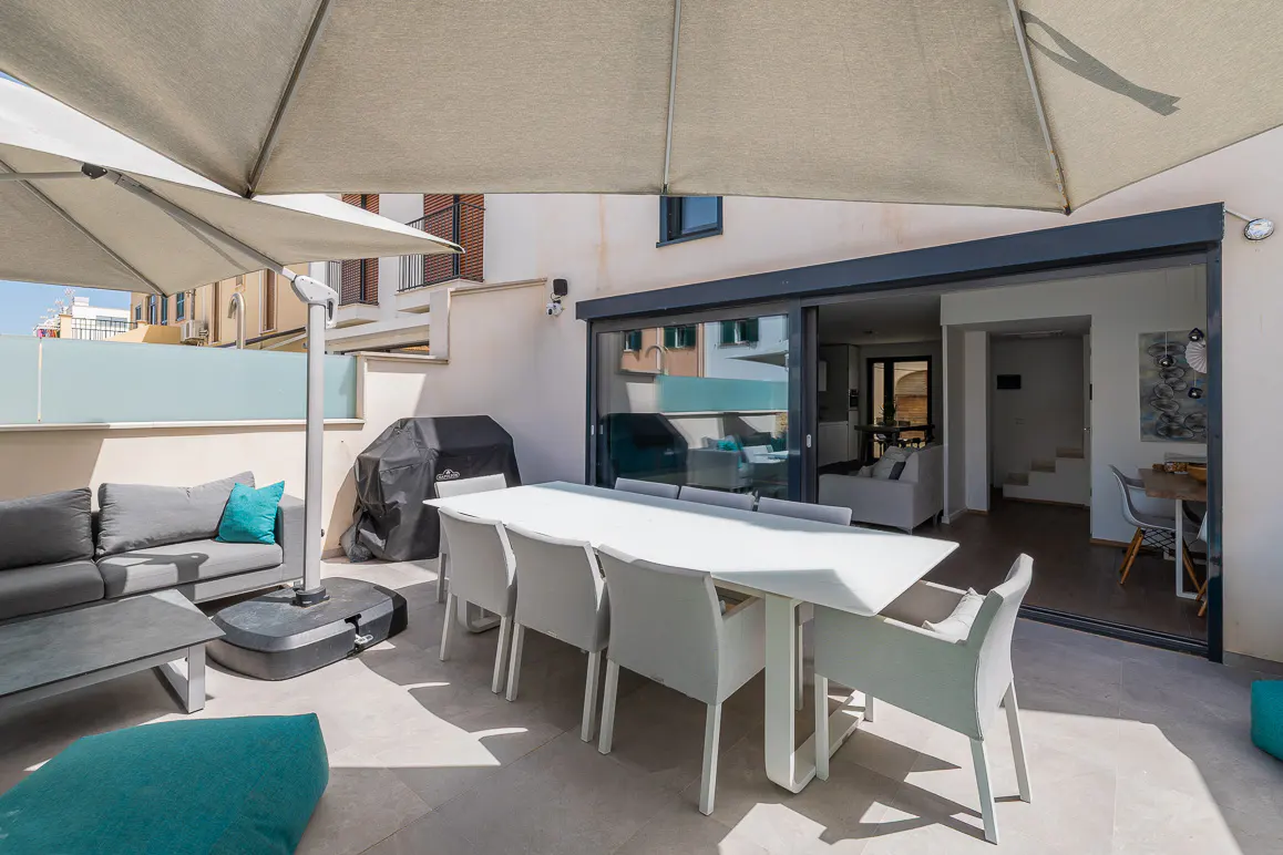 Outdoor patio with a white table, gray chairs, and a gray sofa with a teal pillow under a large umbrella. Sliding glass doors lead to the interior.