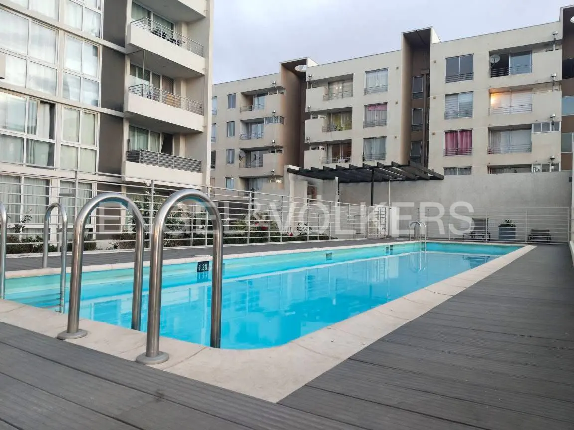 Outdoor pool with blue water and metal ladders, surrounded by gray wood decking and apartment buildings in the background.