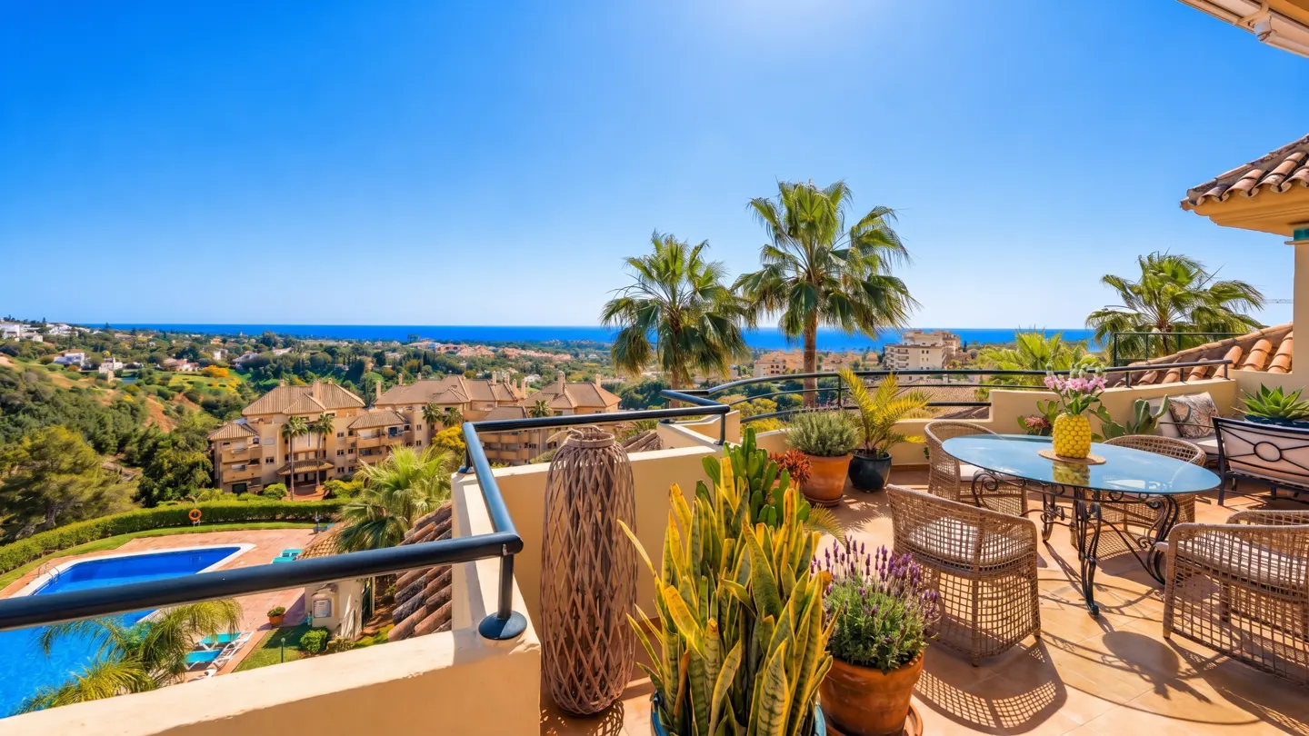 Balcony view with wicker furniture, potted plants, and a blue pool below. Ocean and clear blue sky in the background.
