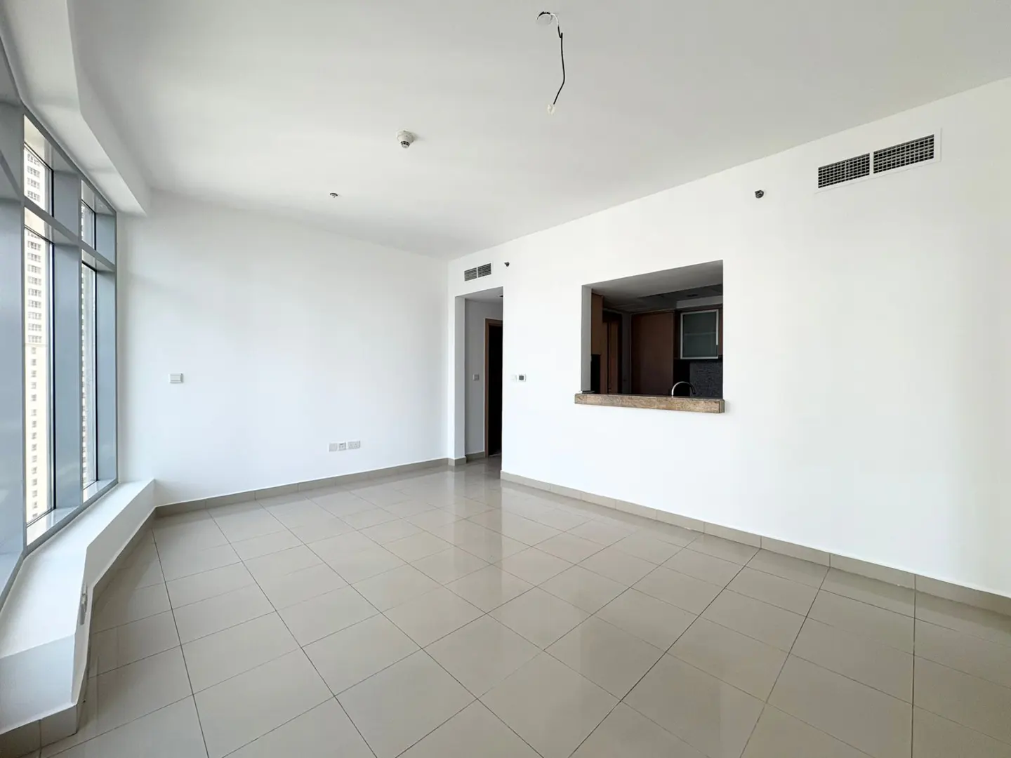 Bright, empty room with white walls, beige tile floor, and large windows. A kitchen pass-through is visible.