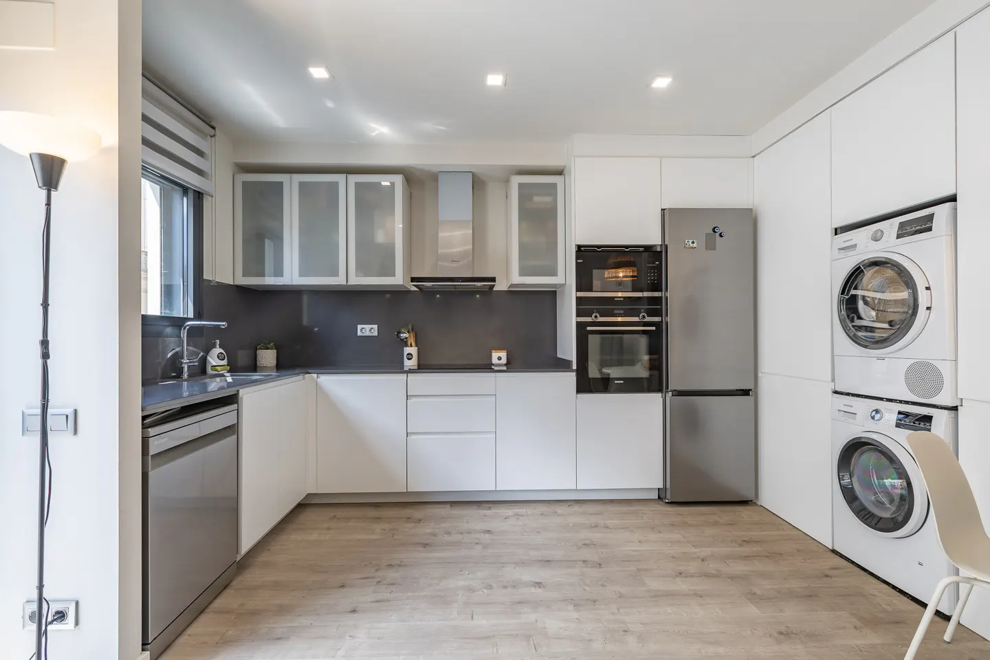 Bright, modern kitchen with white cabinets, stainless steel appliances, and light wood floors. Washer and dryer are stacked on the right.