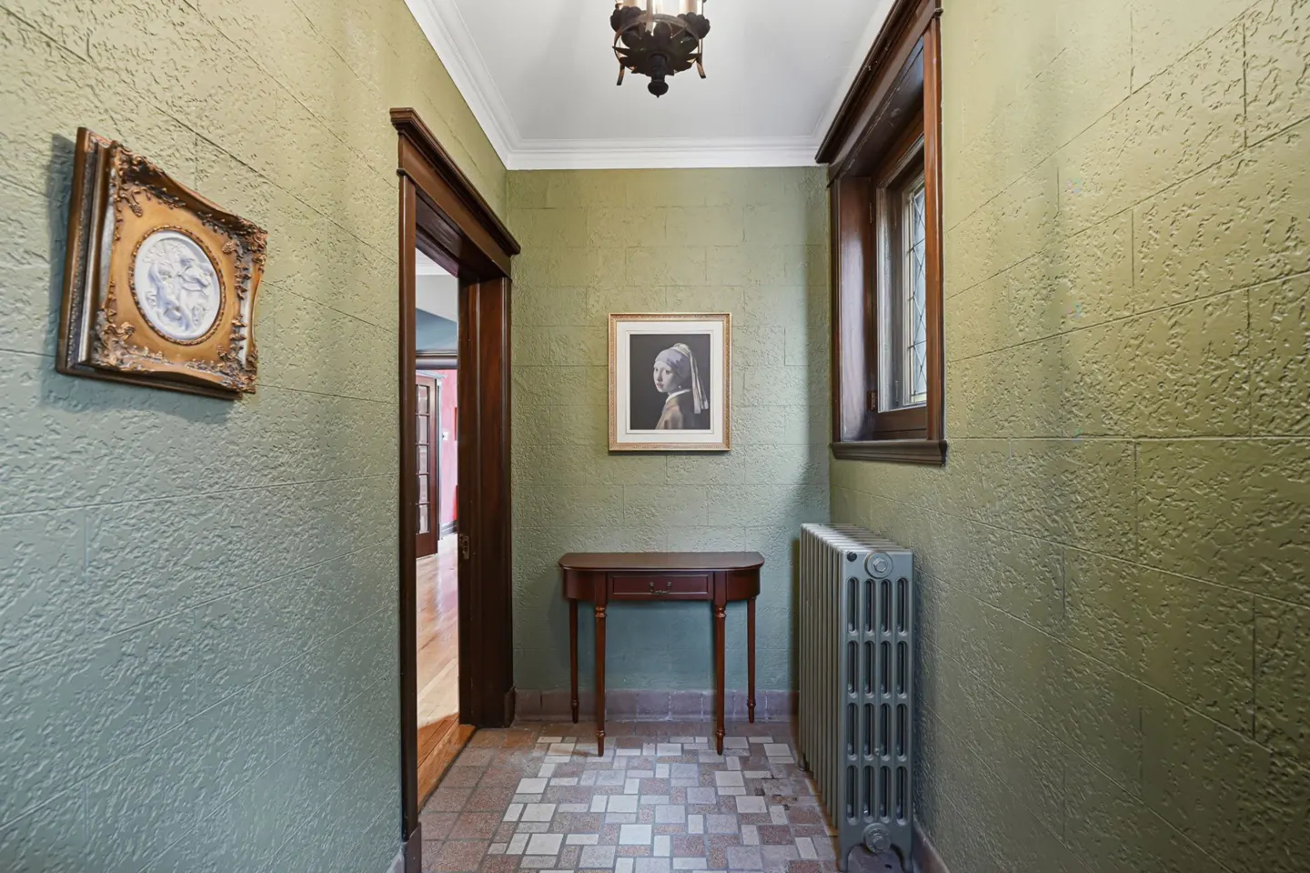 Hallway with green textured walls, tiled floor, and dark wood trim. A table, radiator, and framed art decorate the space.