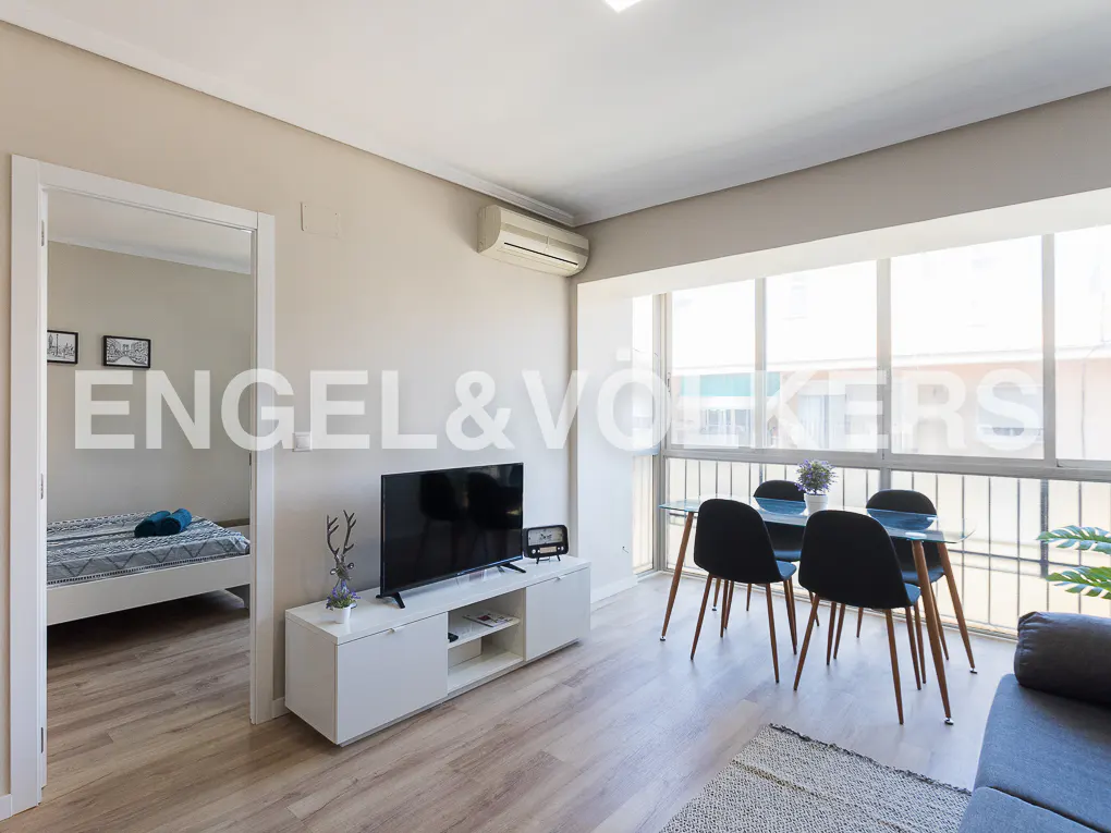 Bright living room with a TV on a white console, a glass dining table with four black chairs, and a view into a bedroom.