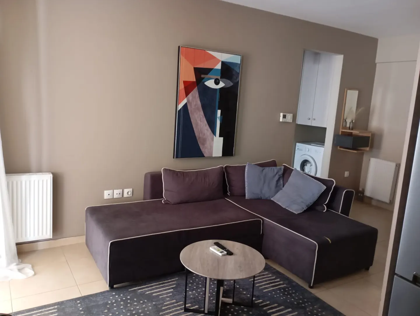 Living room with a brown L-shaped sofa, a round coffee table, and an abstract painting on a beige wall. A washing machine is visible in the background.