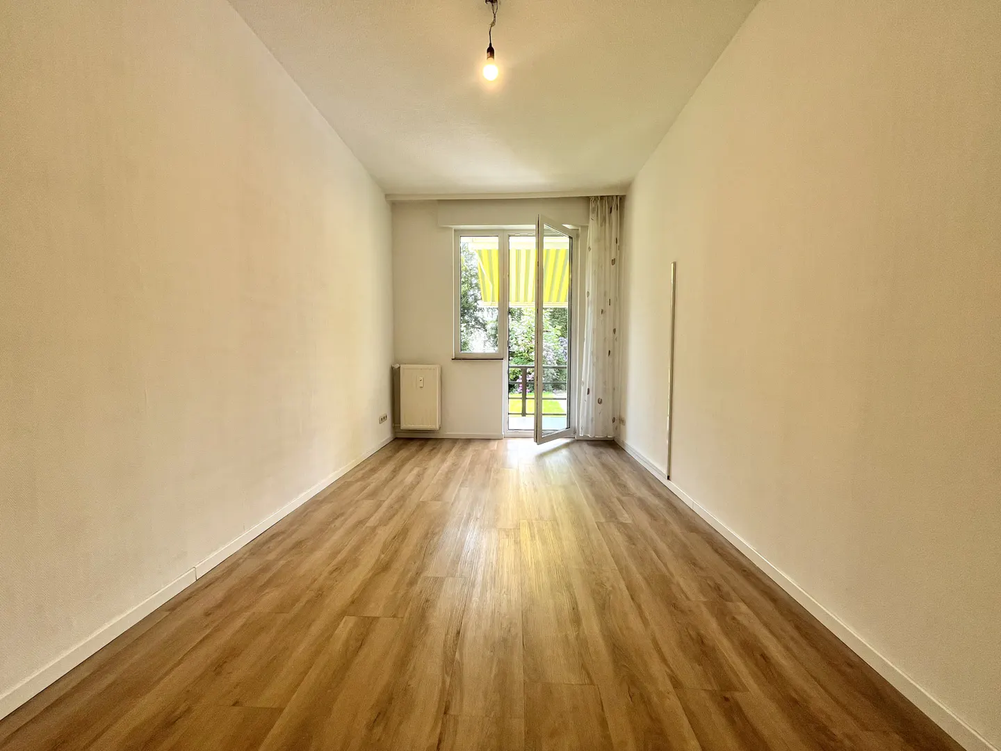 Empty room with light wood floors, white walls, a single pendant light, and a door leading to a balcony with a yellow awning.
