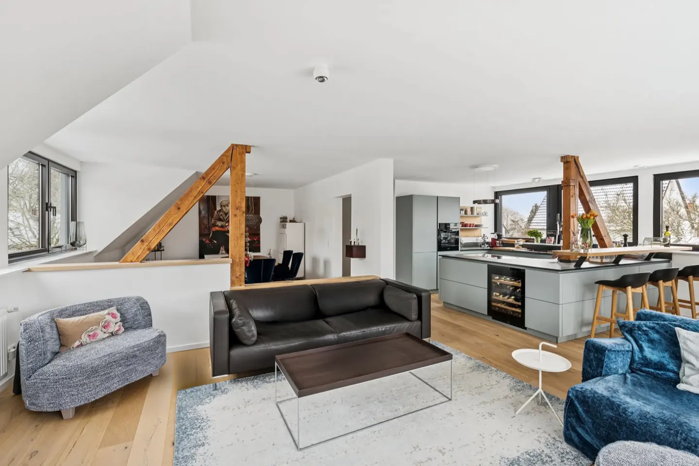 Open-concept living space with a black leather sofa, blue loveseat, and gray kitchen island with bar stools. Wooden beams add character.
