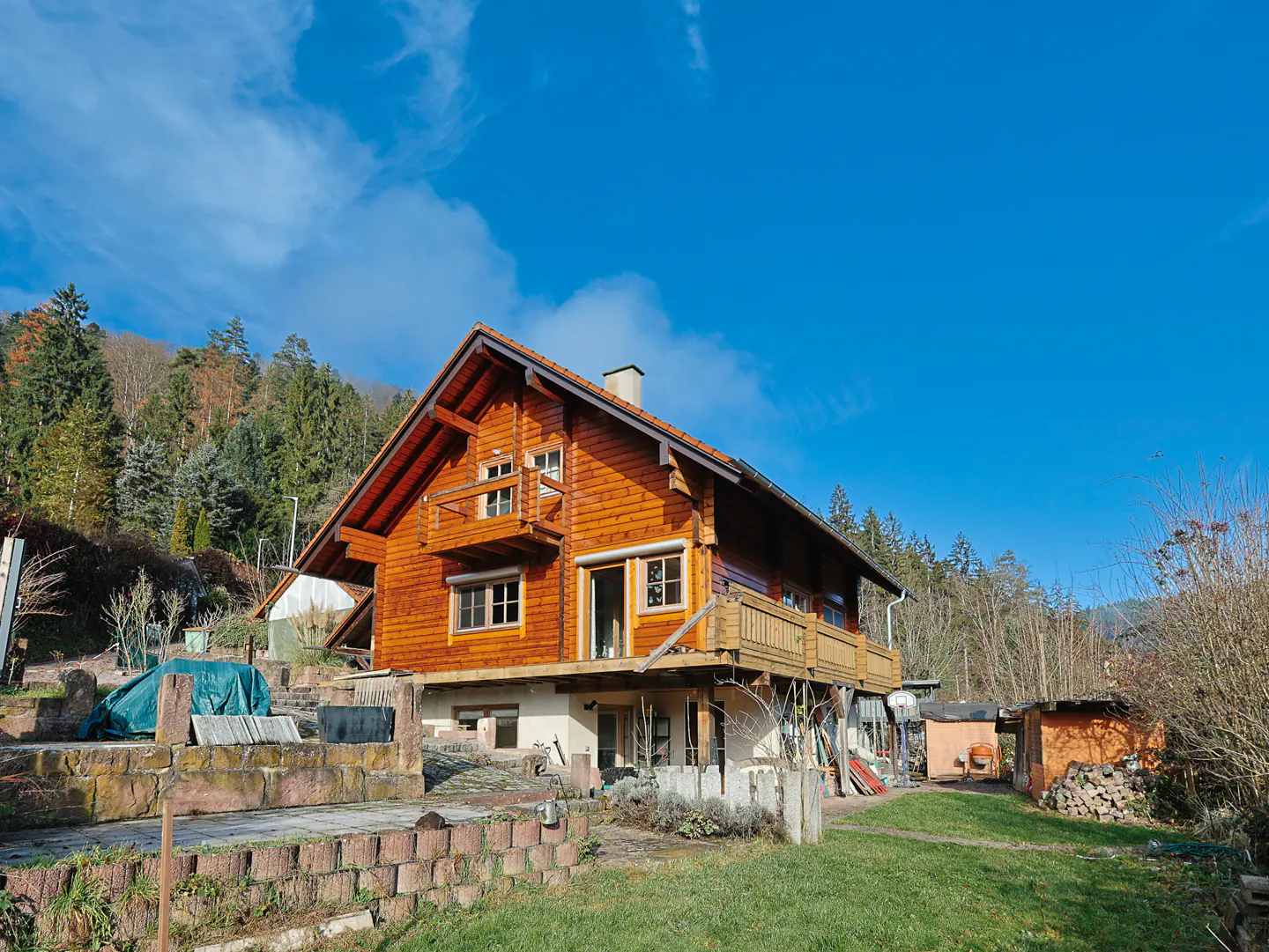 A two-story wooden house with balconies, set against a backdrop of trees and a clear blue sky.