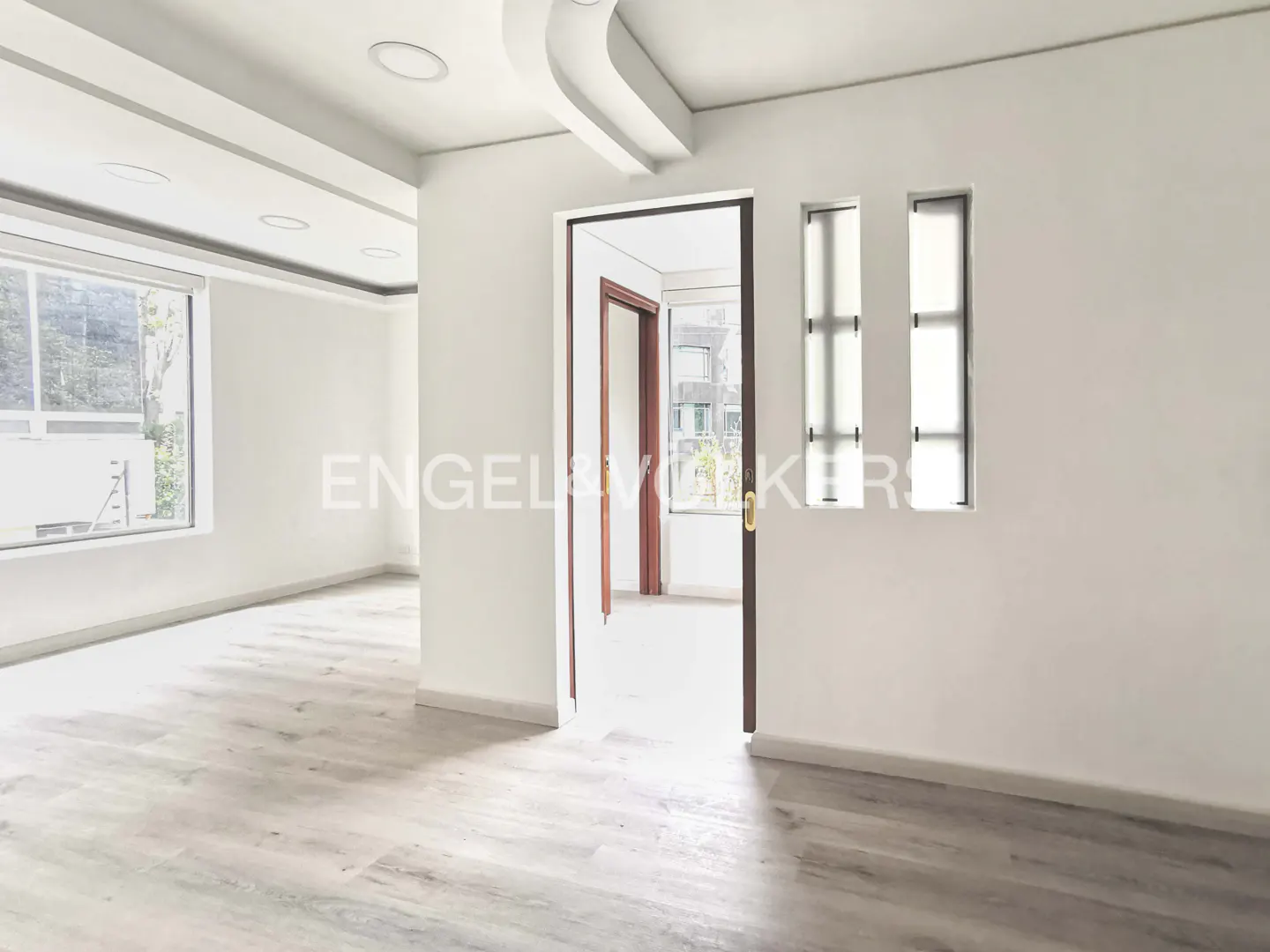 Bright, empty room with white walls, light wood floors, and an open doorway to another room with a window.
