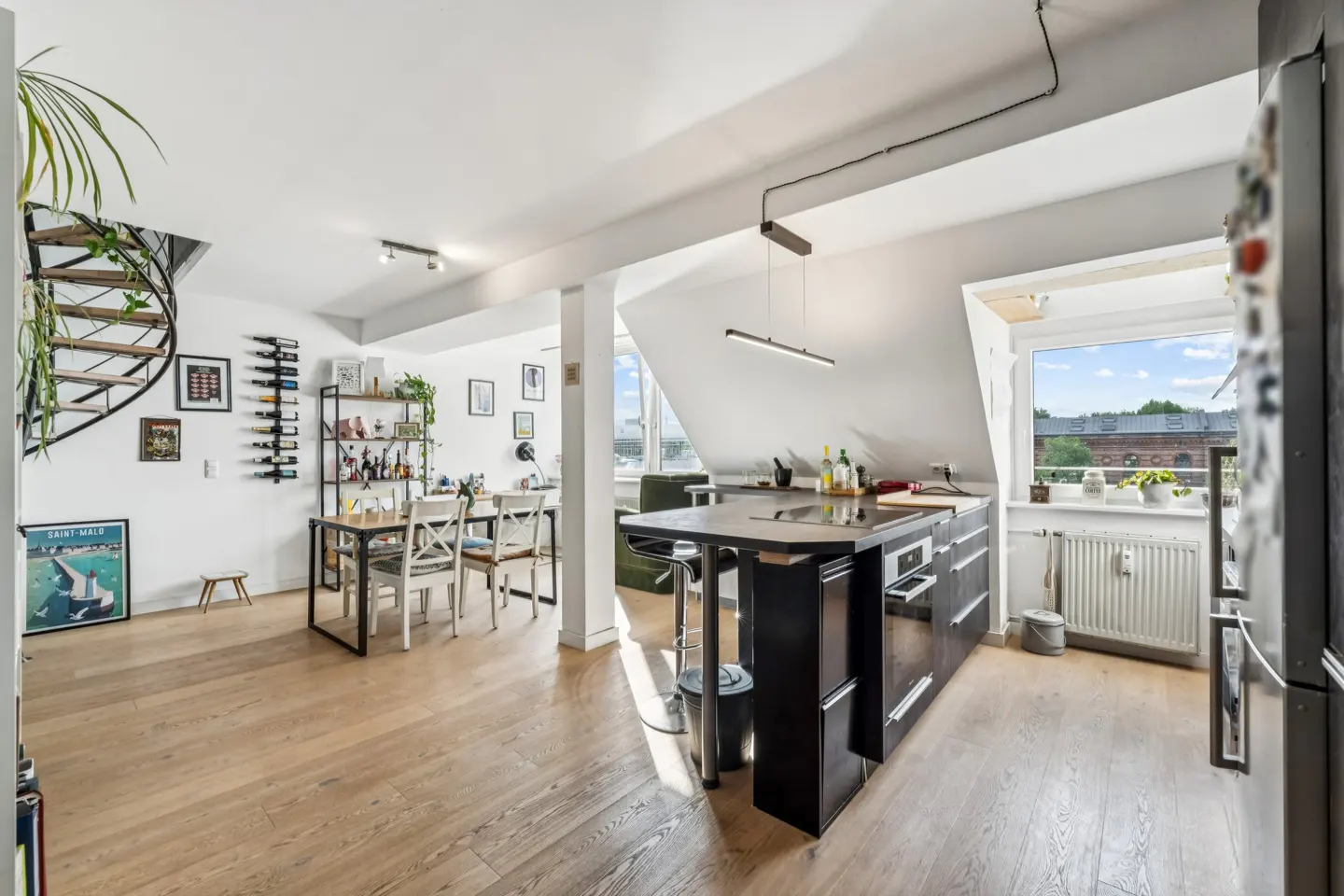 Bright, open-concept apartment with wood floors, a black kitchen island, dining table, and spiral staircase. Sunlight streams through the window.