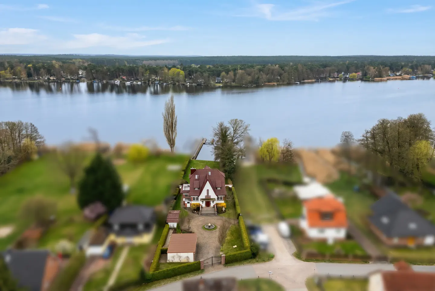 Aerial view of a red-roofed house with a long driveway, green lawn, and a lake in the background.