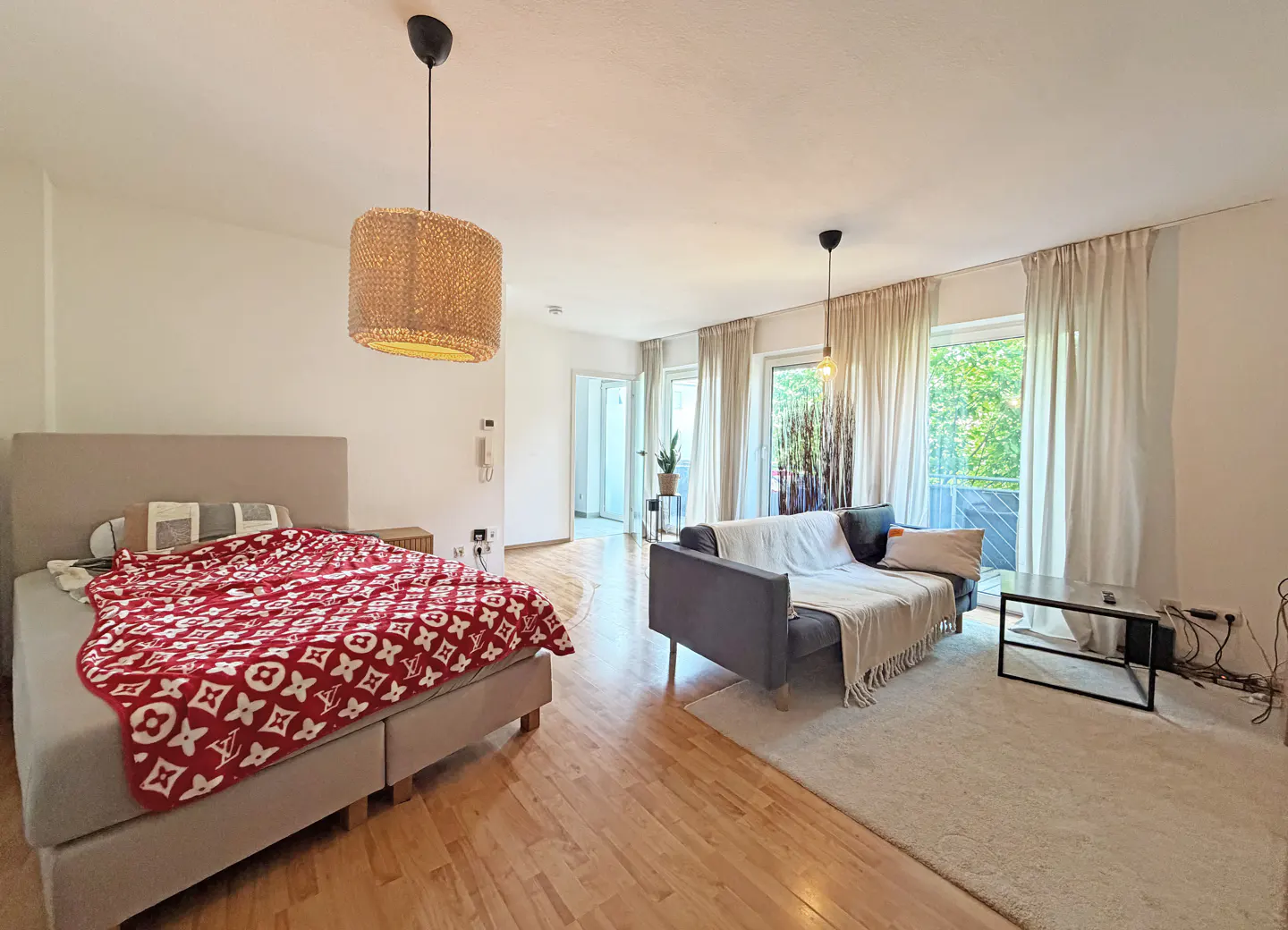 Bright studio apartment with a bed featuring a red patterned throw, a grey sofa, wooden floors, and large windows.