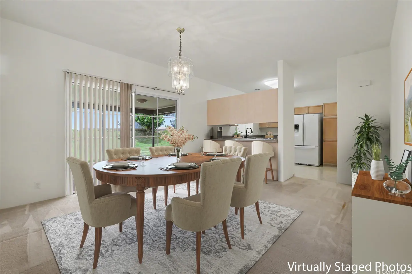 Dining area with oval wood table, beige chairs, and crystal chandelier. Kitchen with bar stools and white appliances in the background.