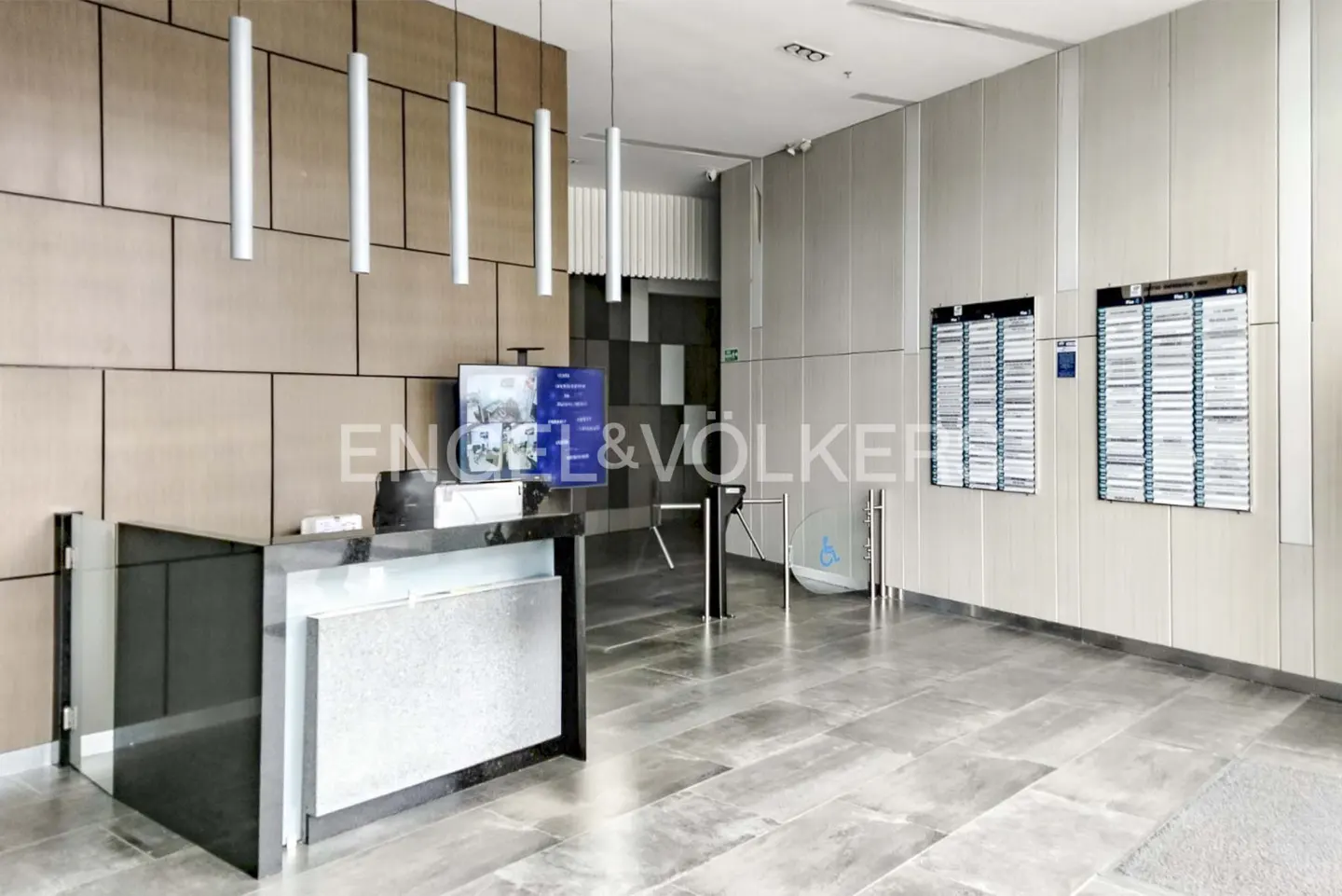 Modern office lobby with a black reception desk, turnstiles, and light wood paneling. Engel & Völkers logo on a screen.