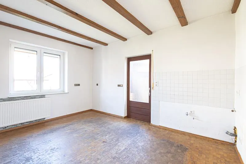 Empty room with white walls, wood floors, and exposed beams. A window and door are visible. The room appears to be under renovation.