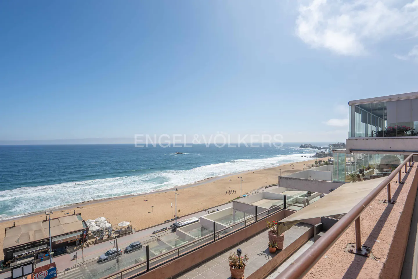 Ocean view from a balcony. Blue water meets a sandy beach under a clear sky. Buildings and a street line the shore.