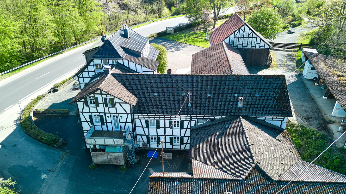 Aerial view of a half-timbered house with black and white facade, dark tiled roofs, and green trees in the background.